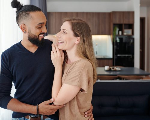 Happy young mixed-race couple in love hugging and looking at each other when standing in studio apartment